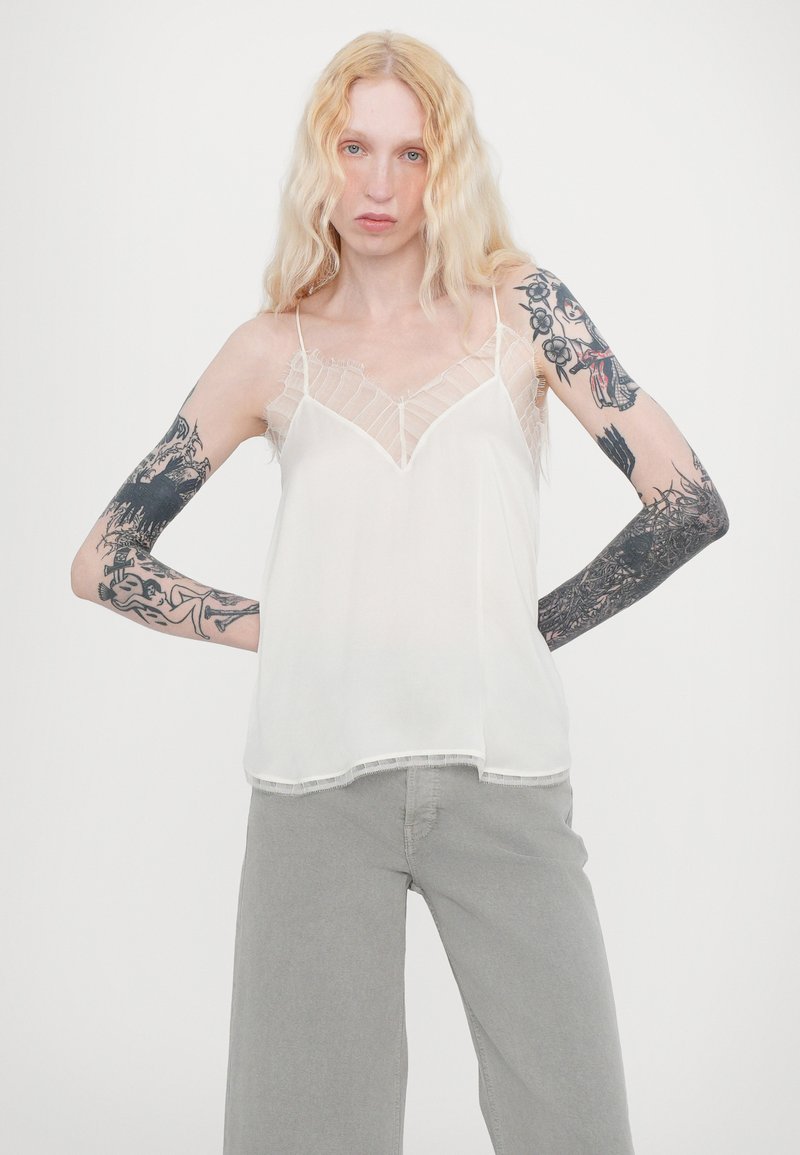 White sleeveless top with sheer lace panel and v-neckline. Lightweight fabric, loose fit, and frayed trim detail at hem. Styled with gray pants.