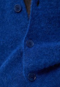 Niebieska tkanina filcowa o teksturowanej powierzchni, z zapięciem na guzik wykonanym z ciemnego plastiku. Skup się na detalu guzika i fakturze materiału.