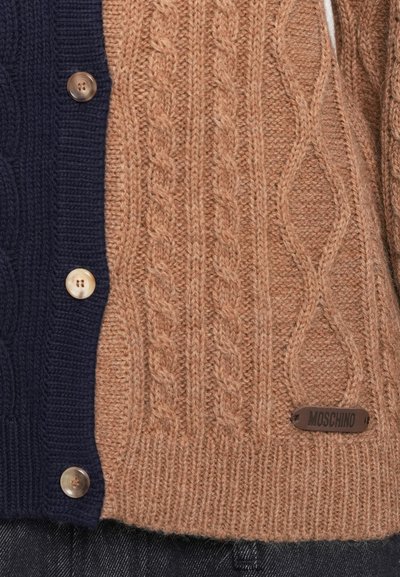 Neulecardigan, jossa on vaalea puuvilla- ja merinovillakudos toisella puolella ja tummansininen toisella puolella. Pyöreät napit ja metallinen Moschino-logo korostavat tyyliä.