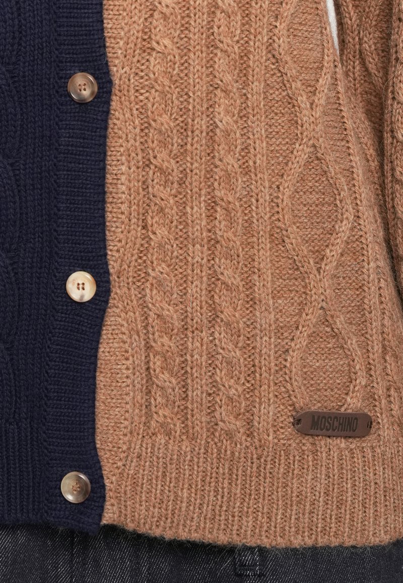 Cardigan lavorato a maglia con un motivo a treccia color beige da un lato e blu navy dall'altro. Bottoni rotondi e un logo Moschino in metallo come dettaglio.