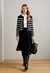 Gilet rayé noir et blanc, blouse blanche, jupe plissée noire, collants noirs, bottes noires jusqu'aux genoux, portant un sac à main noir texturé.