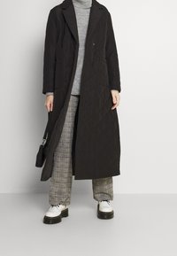 Dlouhý prošívaný kabát tmavě hnědé barvy s klopovým límcem, předními kapsami a délkou ke kolenům, kombinovaný se šedým svetrem a kostkovanými kalhotami.
