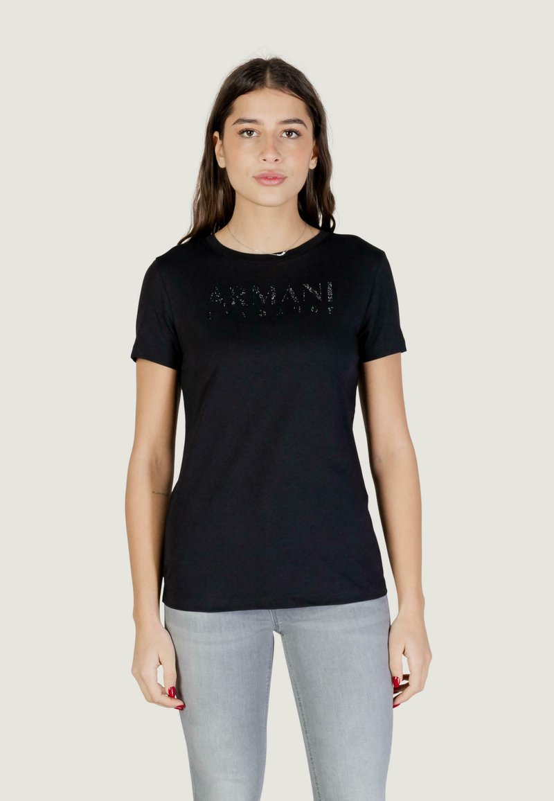 Jonge vrouw met lang bruin haar, gekleed in een zwart Armani T-shirt en lichtgrijze spijkerbroek, staand tegen een effen lichte achtergrond.