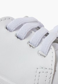 Sneaker bianco con tomaia in materiale sintetico texturizzato, dotata di lacci bianchi e occhielli in metallo. Primo piano dei dettagli cuciti vicino all'area dei lacci.