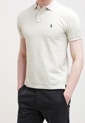 Mand iført en lys beige kortærmet polo med krave og mørke bukser, hænderne i lommerne, stående mod en ensartet baggrund.