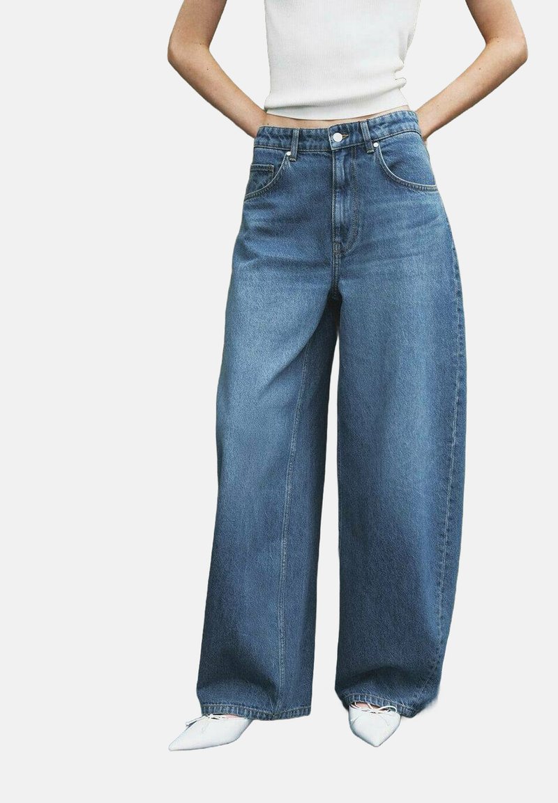 Persona che indossa jeans blu a vita alta e gamba larga, top corto bianco e scarpe bianche a punta, con le mani casualmente nelle tasche.