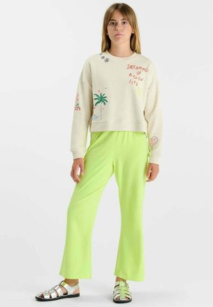 Jonge vrouw staat en draagt een crèmekleurige trui met kleurrijke prints en tekst, neon groene broek en zilveren sandalen met bandjes, neutrale achtergrond.