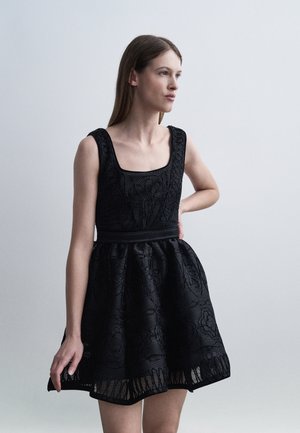 Ung kvinde i en ærmeløs sort blondekjole med tætsiddende krop og udvidet nederdel, stående med den ene hånd på hoften.