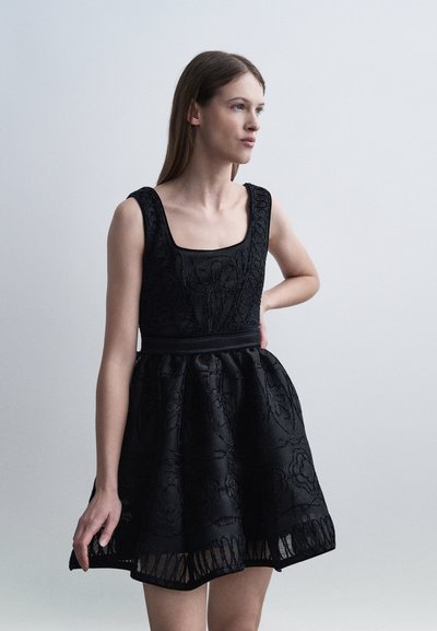 Jeune femme en robe noire sans manches en dentelle avec un corsage ajusté et une jupe évasée, debout avec une main sur la hanche.