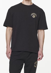 Černé bavlněné tričko s volným střihem, na levé hrudi se zlato-žlutým logem "PEGADOR"; krátké rukávy a kulatý výstřih.