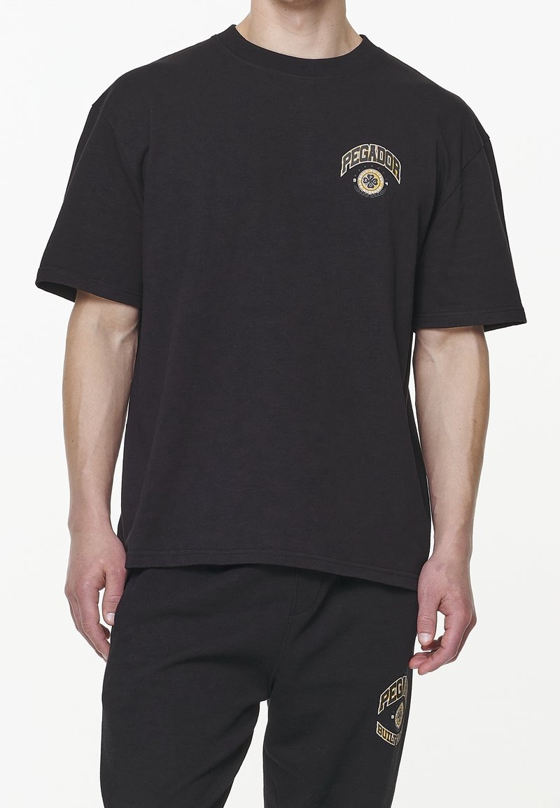 Černé bavlněné tričko s volným střihem, na levé hrudi se zlato-žlutým logem "PEGADOR"; krátké rukávy a kulatý výstřih.