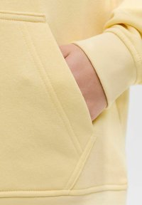 Sweat à capuche jaune en coton avec une poche kangourou. La texture est lisse, avec des poignets côtelés et un ourlet droit. Les détails de couture sont visibles.