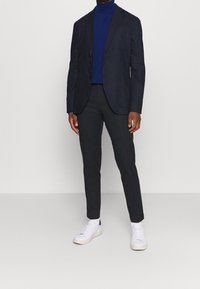 Marineblazer mit zwei Vordertaschen, getragen über einem blauen Rollkragenpullover. Schmale dunkelgraue Hose und weiße Sneakers vervollständigen das Outfit. Minimalistisches Design.