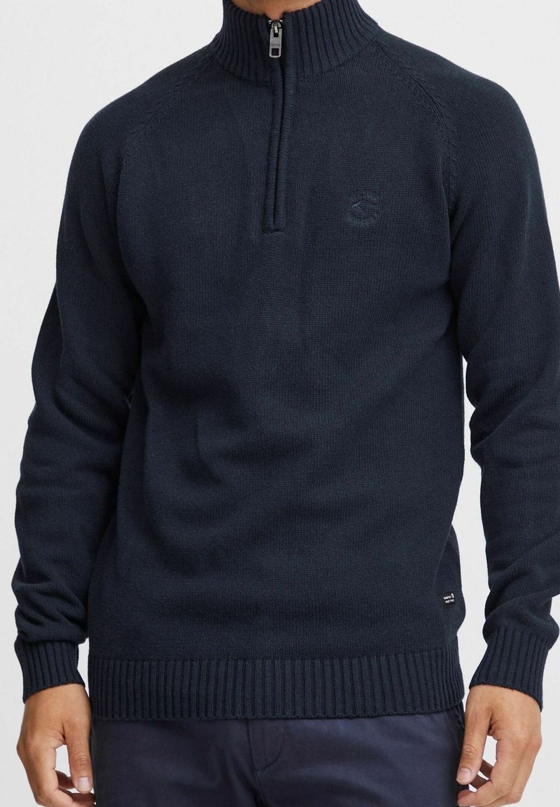 Pull en maille bleu marine avec un col montant, une fermeture à demi-zip, des poignets et un ourlet côtelés. Présente un logo discret sur la poitrine.