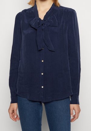 Marinblus med knytkrage, långa ärmar och knapplist. Har en mjuk textur och diskret glans, accentuerad med guldfärgade knappar.