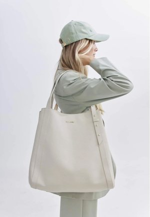 Beige draagtas met een gestructureerde afwerking, lange handvatten, gouden accenten en decoratieve knopen. G gedragen met een lichtgroene outfit en een pet.