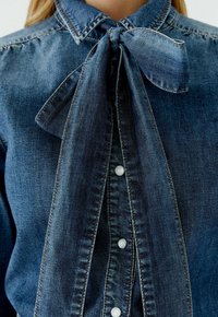 Jeansbluse in Dunkelblau mit einer Schleifenkragen, ausgestattet mit einer Knopfleiste mit perlmuttartigen Knöpfen und sichtbaren Steppnähten.