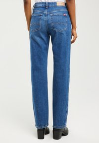 Widok z tyłu osoby ubranej w niebieskie dżinsy o prostych nogawkach i czarne buty na obcasie na jednolitym tle.