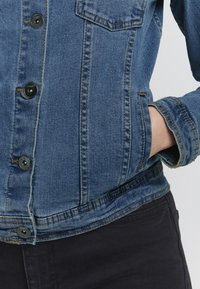 Jeans jakna v svetlo modri barvi s črnimi gumbi, ki ima sprednji žep, robustno šivanje in nekoliko skrajšan rob.