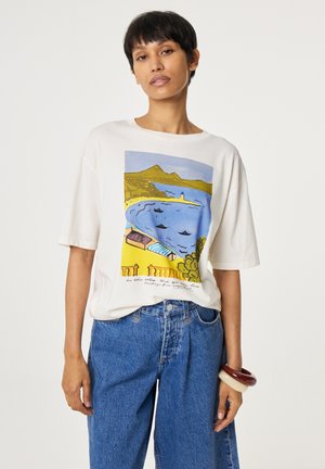 Frau mit kurzen schwarzen Haaren, die ein weißes T-Shirt mit einem bunten Küstenlandschaftsprint und blaue Jeans trägt, posiert vor einem weißen Hintergrund.