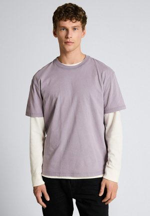 Młody mężczyzna o kręconych włosach, ubrany w lawendową koszulę z krótkim rękawem na białym topie z długim rękawem oraz czarne spodnie, stojący na jednolitym tle.