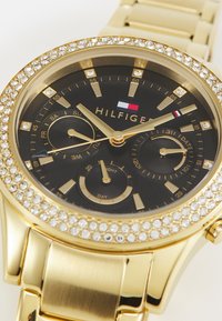 Montre-bracelet dorée avec un cadran noir, ornements en cristal autour de la lunette, et deux sous-cadrans pour les indicateurs de date et de jour.