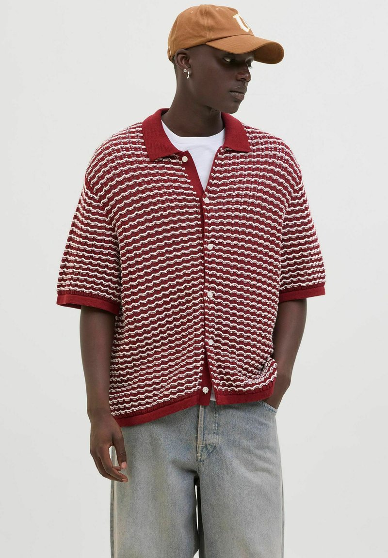 Cardigan en tricot à manches courtes rayé rouge et blanc avec un col et une fermeture à boutons, porté par-dessus un t-shirt blanc et associé à un pantalon en denim clair.
