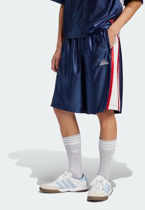 Osoba w granatowych, koszykarskich spodenkach Adidas z czerwono-białymi paskami po bokach, szarych skarpetkach za kostkę Adidas i białych trampkach z niebieskimi paskami.