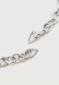 Κοντινό πλάνο ενός ασημένιου βραχιολιού αλυσίδας με καρδιοειδείς συνδέσμους, ένας σύνδεσμος ανοιγμένος, σε απλό φωτεινό φόντο.