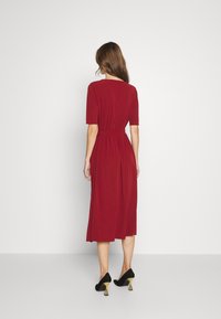 Rotes, knielanges Kleid mit kurzen Ärmeln, Taillengürtel, fließendem Stoff und glatter Textur. Getragen mit schwarzen High Heels mit goldenen Akzenten.