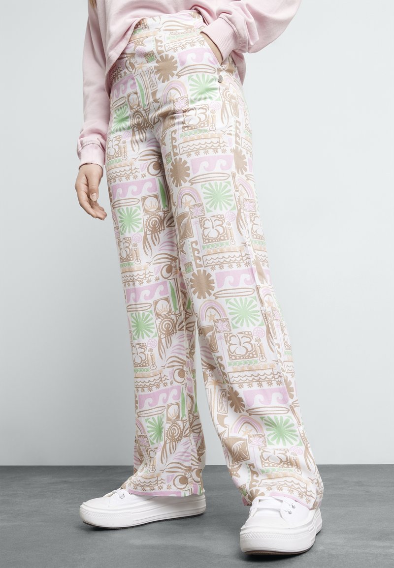 Personne portant un pantalon taille haute avec des motifs floraux et géométriques abstraits dans des tons pastel, associé à des baskets blanches et un sweat-shirt rose.