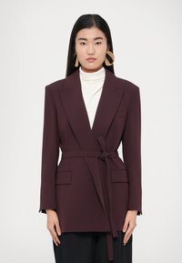 Bordo színű blézer övvel, bevágott hajtókákkal és két első zsebbel, fehér garbó fölött rétegezve. Sima anyagtextúra.