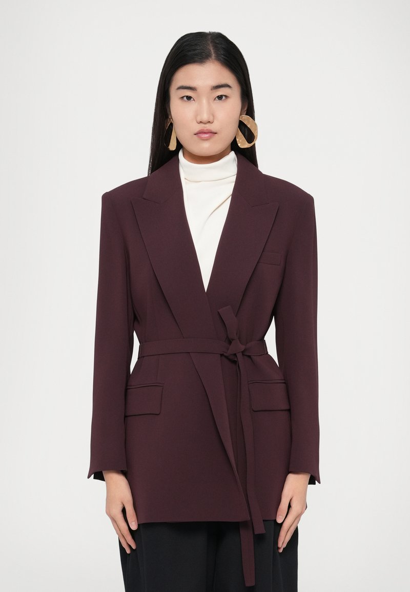 Bordo színű blézer övvel, bevágott hajtókákkal és két első zsebbel, fehér garbó fölött rétegezve. Sima anyagtextúra.