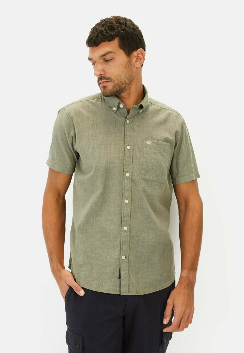 Kortærmet olivengrøn skjorte med knapper og lomme, struktureret stof og et subtilt mønster, båret sammen med sorte cargo shorts.
