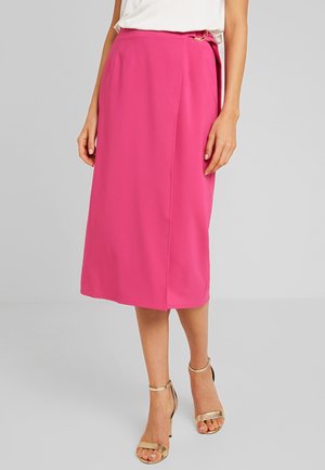 Femme portant une jupe midi rose vif avec une boucle sur le côté, un haut blanc et des sandales à talons dorés, debout sur un sol gris.