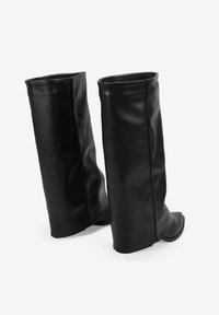 Un par de botas de cuero negro hasta la rodilla con tacones ocultos, de pie en un fondo blanco.