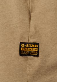 Tejido color marrón claro con un acabado texturizado; presenta una etiqueta negra con texto amarillo que dice "G-STAR ORIGINALS RAW DENIM."