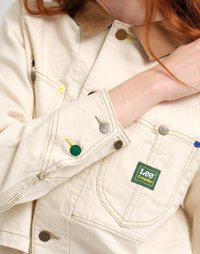 Κρέμα καμβάς μπουφάν με καφέ γιακά, μεταλλικά κουμπιά και πολύχρωμες ραφές. Διαθέτει πράσινη ετικέτα λογότυπου στην τσέπη του στήθους.