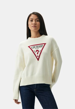 Maglione color crema con orlo a coste, caratterizzato da un logo triangolare rosso prominente con la scritta "GUESS® U.S.A." in testo nero.