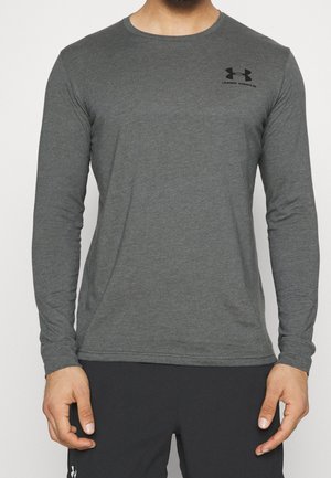Mann trägt ein eng anliegendes dunkelgraues Langarmshirt mit schwarzem Under Armour-Logo auf der Brust und schwarze Sporthose.