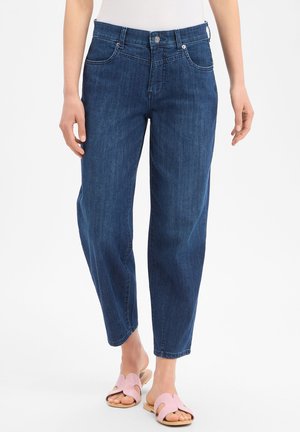 Vrouw met donkerblauwe jeans en roze open teen slippers, staand tegen een witte achtergrond.
