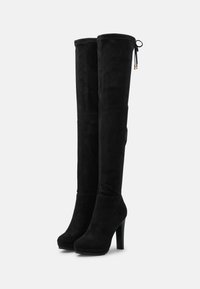 Botas negras hasta la rodilla con un acabado de ante suave, tacones de aguja altos y detalle decorativo de lazo en la parte superior. Textura lisa y diseño elegante.
