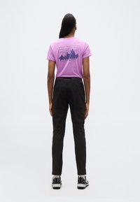 Γυναίκα που στέκεται με την πλάτη, φοράει ένα ροζ t-shirt Patagonia με γραφικό βουνού και κείμενο, μαύρο παντελόνι, λευκές κάλτσες και παπούτσια πεζοπορίας.