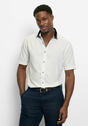 Mann trägt ein weißes Kurzarmhemd mit schwarzem Kragenbesatz und dunkelblaue Hose, steht mit einer Hand in der Hosentasche vor weißem Hintergrund.