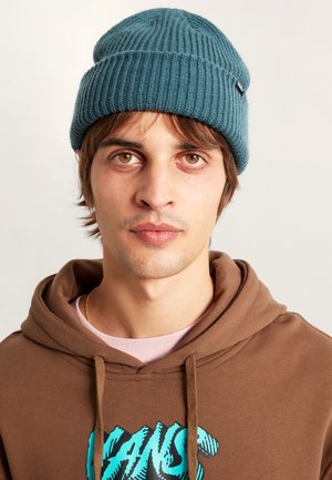 Tyrkysová pletená čepice s vroubkovaným vzorem, vyrobená z vlněné směsi, nošená přes hnědou mikinu s tyrkysovým logem. Má ohrnutý okraj. Snadný a texturovaný design.