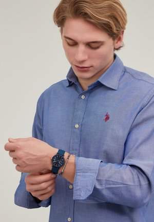 Jeune homme en chemise bleue ajustant une montre bleue sur son poignet gauche, portant un bracelet en argent, regardant sa montre sur un fond uni.