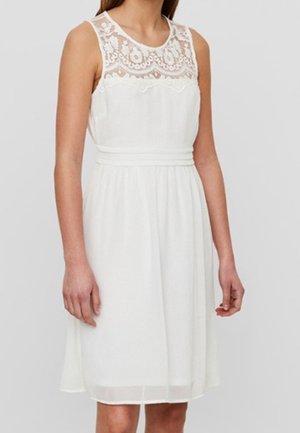 Femme portant une robe blanche sans manches, longueur genou, avec des détails en dentelle sur le haut de la poitrine et une taille ajustée.