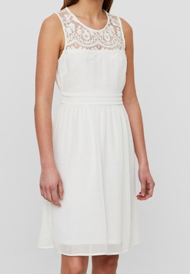Femme portant une robe blanche sans manches, longueur genou, avec des détails en dentelle sur le haut de la poitrine et une taille ajustée.