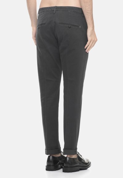 Man in een donkergrijze opgerolde broek en zwarte gepolijste dresschoenen, gezien vanaf de achterkant tegen een witte achtergrond.