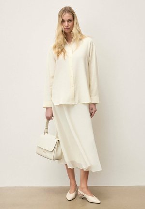 Naine, kes kannab kreemikat nööbitavat pluusi, sobivat midi-seelikut ja slingback-kingi, hoides kreemjat käekotti ühetaustal heledal taustal.